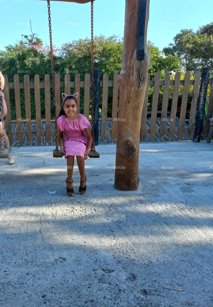 Criança brincando no balanço, usando vestido rosa, arquinho na cabeça com orelhas e sandália preta