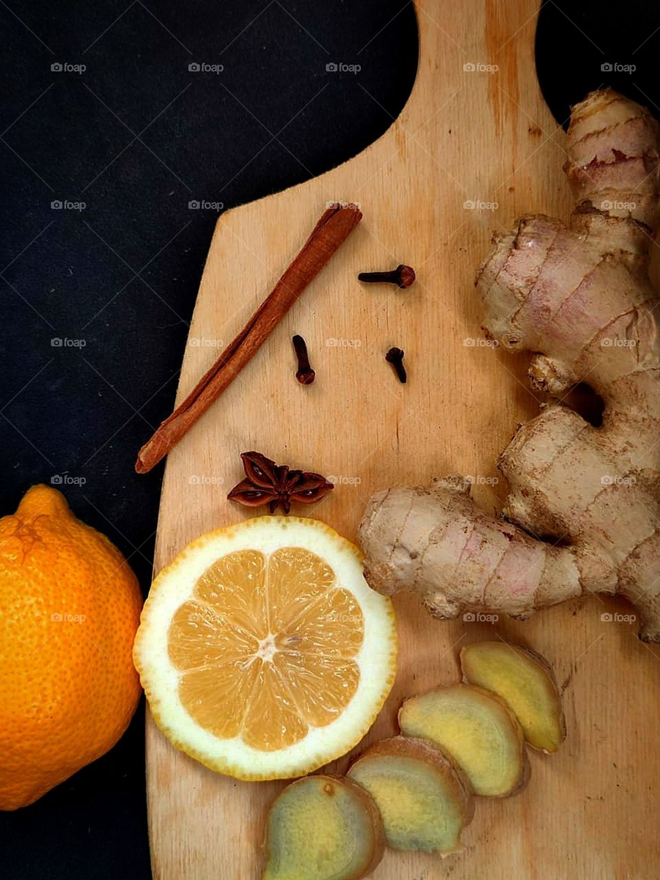 Wooden board containing: sliced ​​lemon, anise, cinnamon stick, ginger root and sliced ​​ginger.  There is a lemon near the board.  Black background