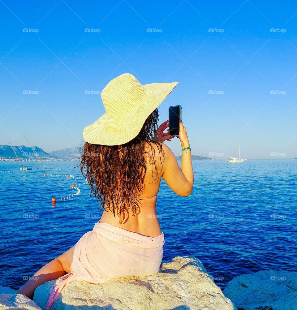 A young woman sits on a rock by sea and enjoys photographing the seascape with a mobile phone