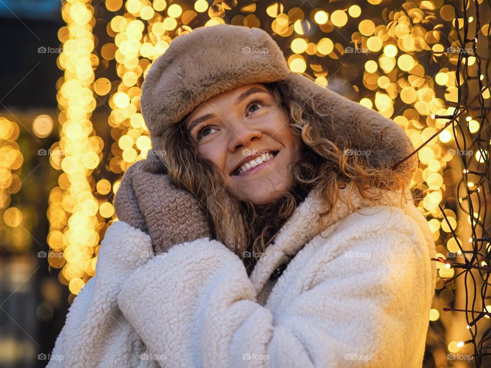 Young beautiful woman with curly hair in fur coat and hat with ear flaps smiling with lights on a background 