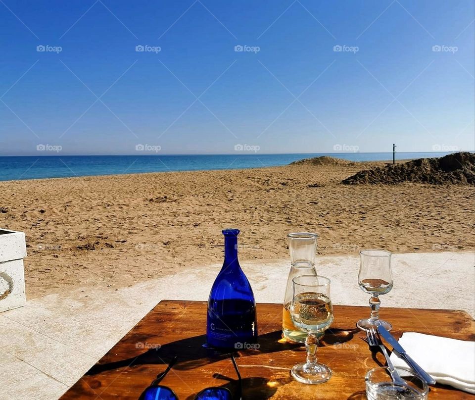 Sea shore. Beach. On the golden sand stands a wooden table for two persons. On the table are two glasses of white wine, a bottle of water, and sunglasses. View of the blue sea, which merges with the blue sky on the horizon