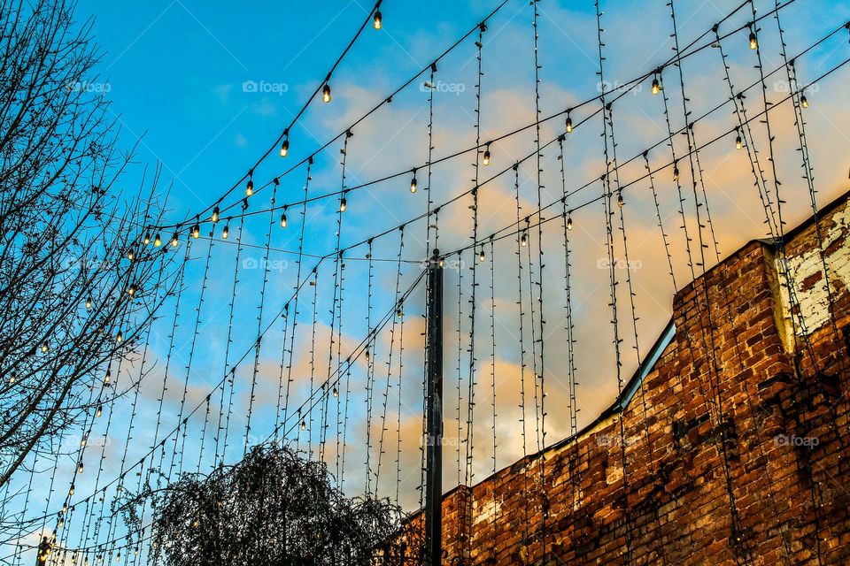 A setting sun creates bright blue and orange hues behind a string light decorated bridge, seen from an angle well below the path of the bridge