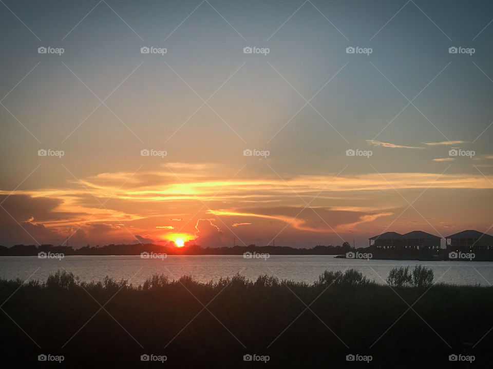 Sun setting over Inlet water with foliage and silhouette of houses on stilts.