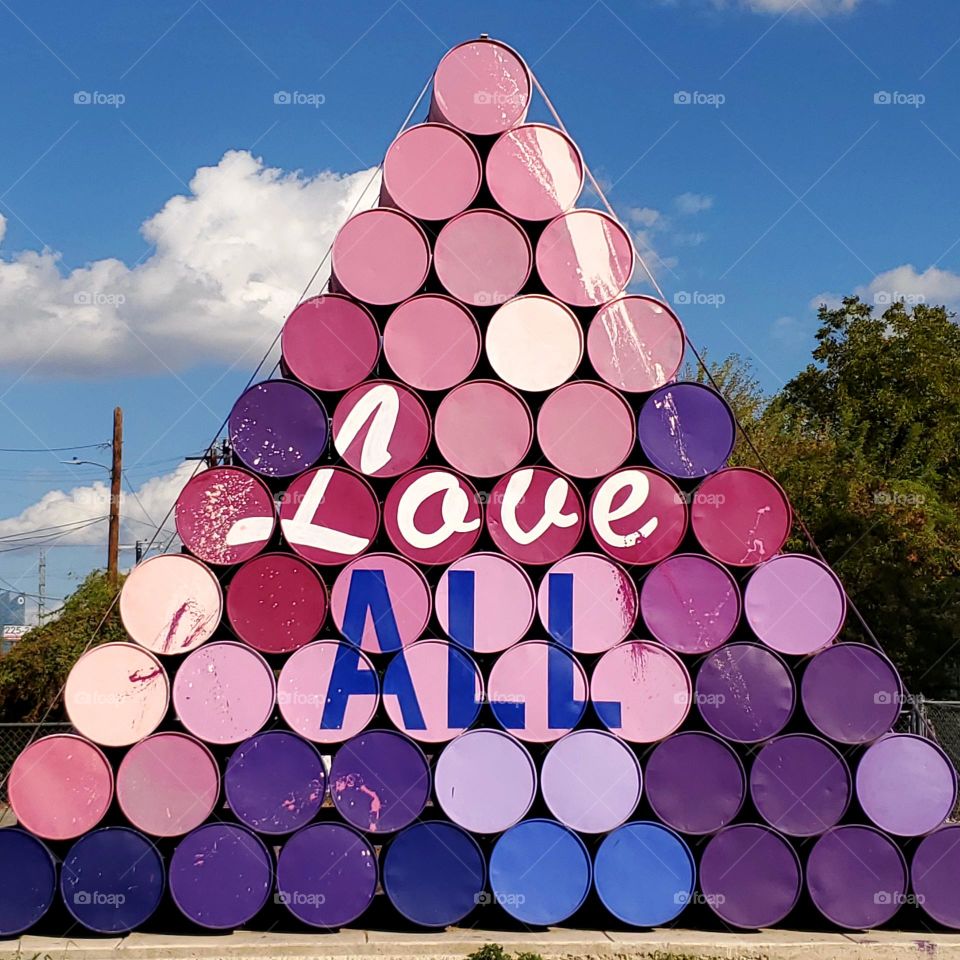 Outdoor urban art made out of colorfully painted stacked barrels in the shape of a triangle with the words Love ALL.