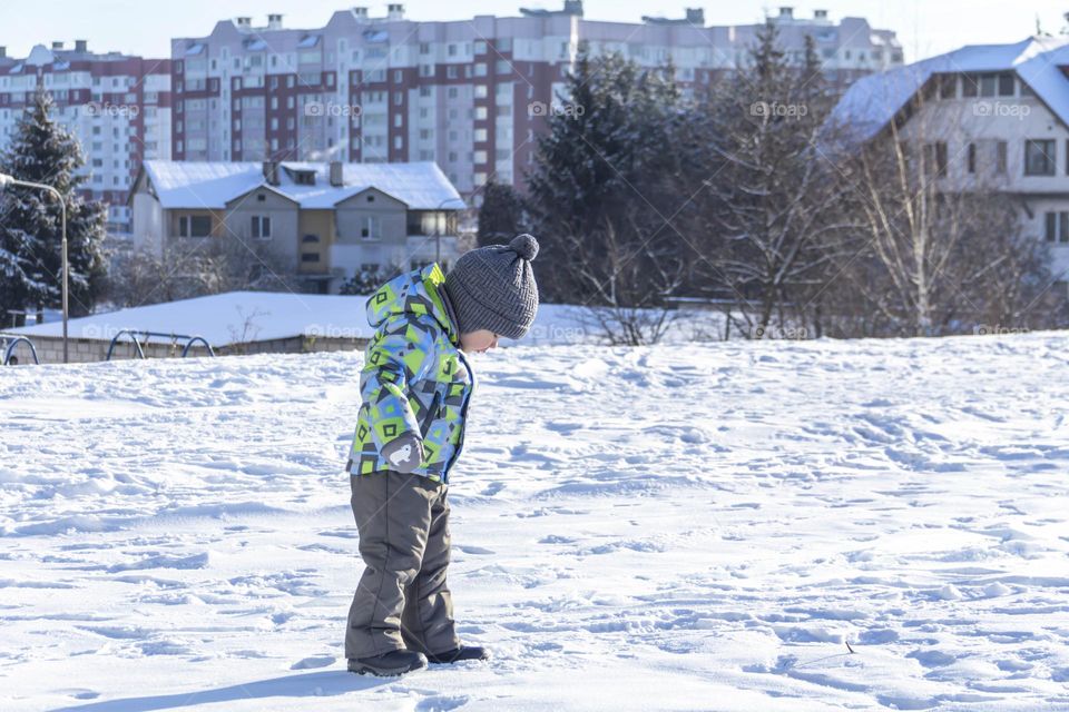 A child with a serious expression on his face in winter clothes jackets, pants, hat and boots in winter on the white snow on the street and in the park in nature plays winter fun.