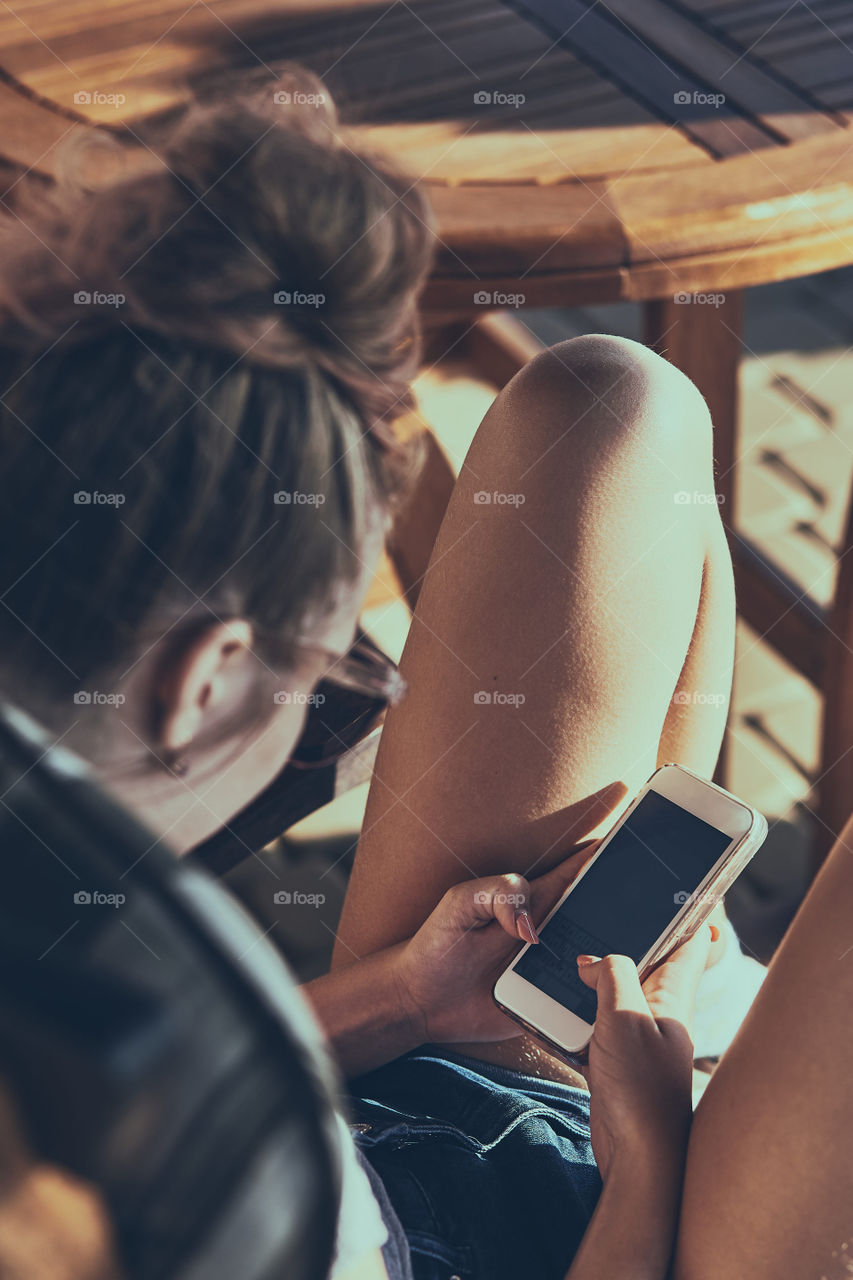 Young woman chatting on a mobile phone sitting in a chair outdoors on patio wearing a sunglasses. Real people, authentic situations
