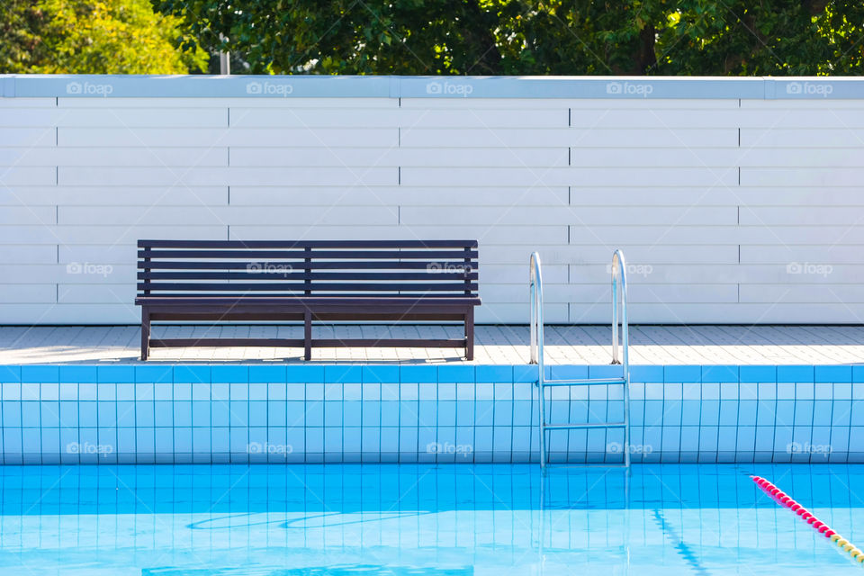 Side of a swimming pool showing the pool, a wall with trees behind it, a bench, and a ladder