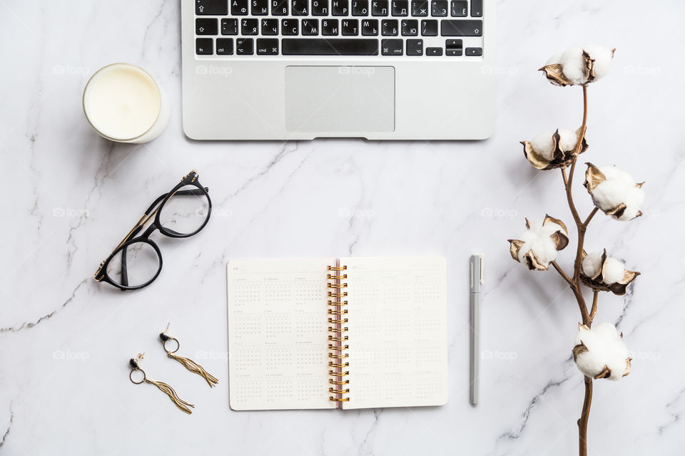 Desktop flatlay items: laptop, calendar, eyeglasses, candle, earrings, pen, cotton flowers lying on marble background 