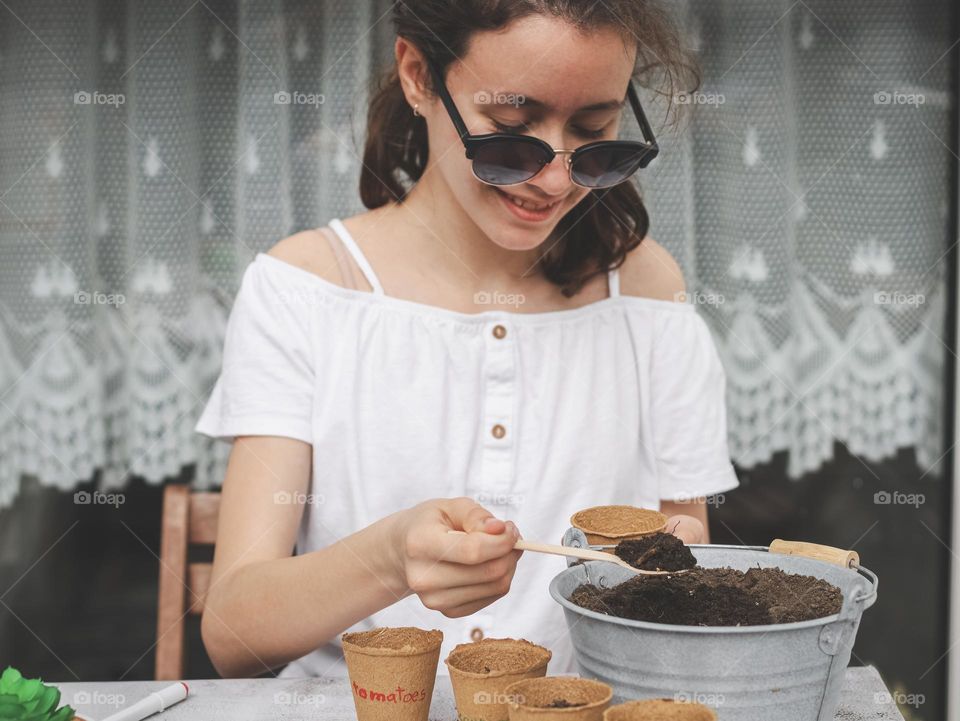 Beautiful caucasian girl teenager brunette in sunglasses with a smile on her face pours black soil into a cardboard craft glass from a zinc bucket with a wooden spoon, sitting at a light stone table against the background of a window in the backyard