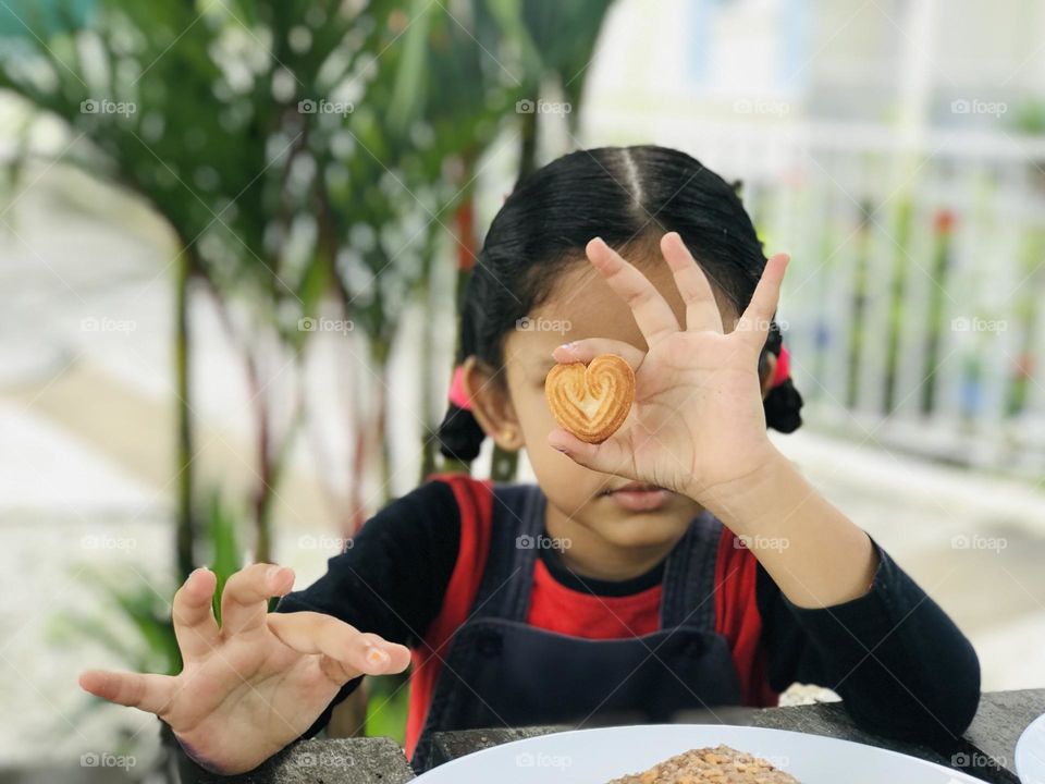 A cute girl holding heart shaped cookie with her fingers and showing towards camera 