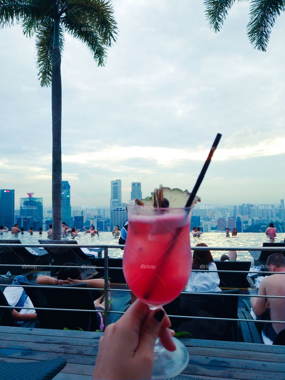 Enjoying a great Singapore Sling at the infinity pool in the Marina Bay Sands Hotel in Singapore. With tons of other people in the adult pool and a timid skyline o in the back.