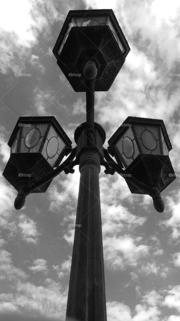 Unique Contrasts In This Black And White Photo. Looking Up Through A Silhouetted Three Light Lightpost With A Sky Background Of Fluffy Clouds.