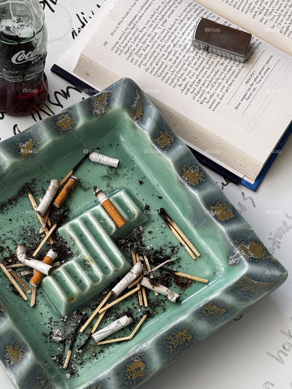 Heath-style turquoise celadon and bronze accented large ceramic ashtray with cigarette butts and matches with Coke bottle and “How to Win Friends and Influence People” with Zippo lighter