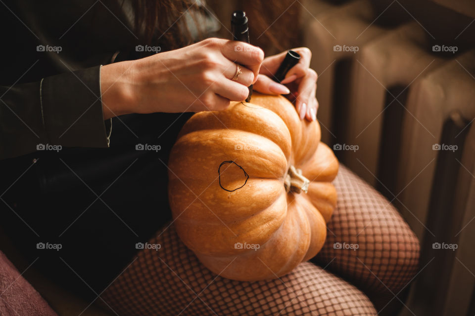 A cute young girl is sitting at a table in the living room painting her face on a big pumpkin for Halloween. Halloween holiday and family lifestyle background.