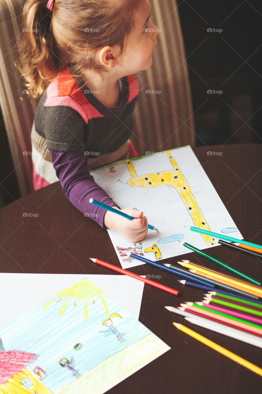 Little girl drawing a colorful pictures of giraffe and playing children using pencil crayons sitting at table indoors. Shot from above