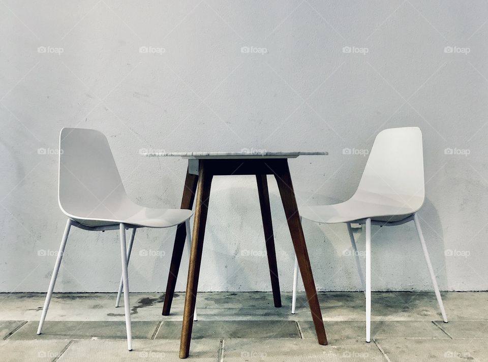 Pair of white cafe chairs and wooden cafe table on concrete floor against white exterior wall
