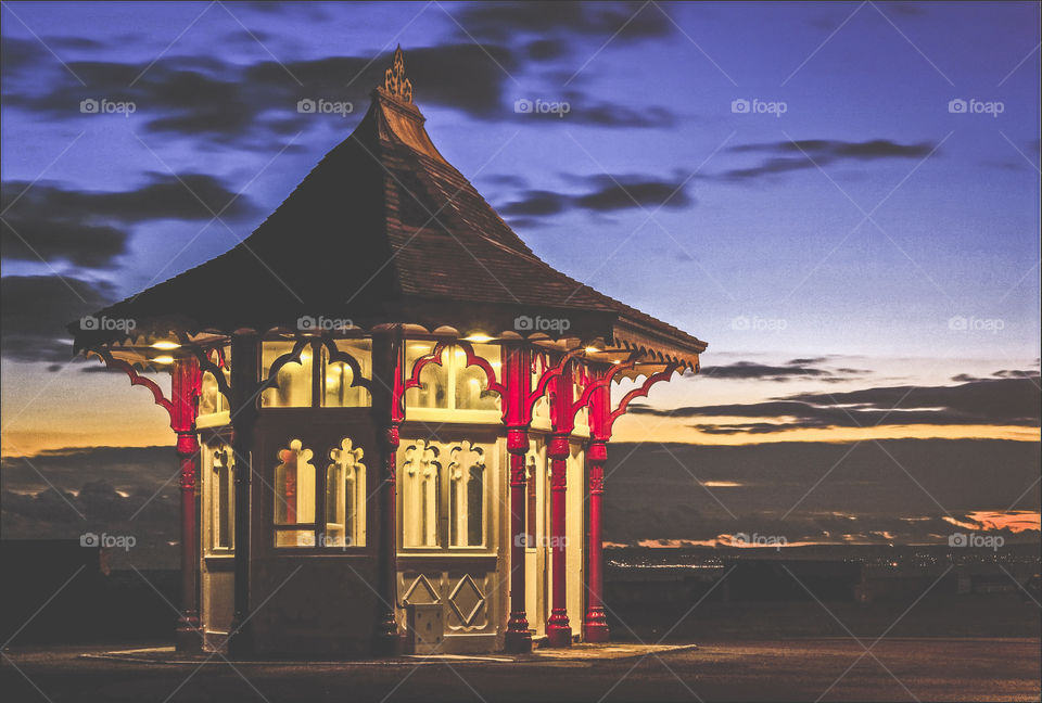 A Victorian seafront shelter at Bexhill, lit up against the evening sky, lights from nearby Eastbourne can be seen in the far distance