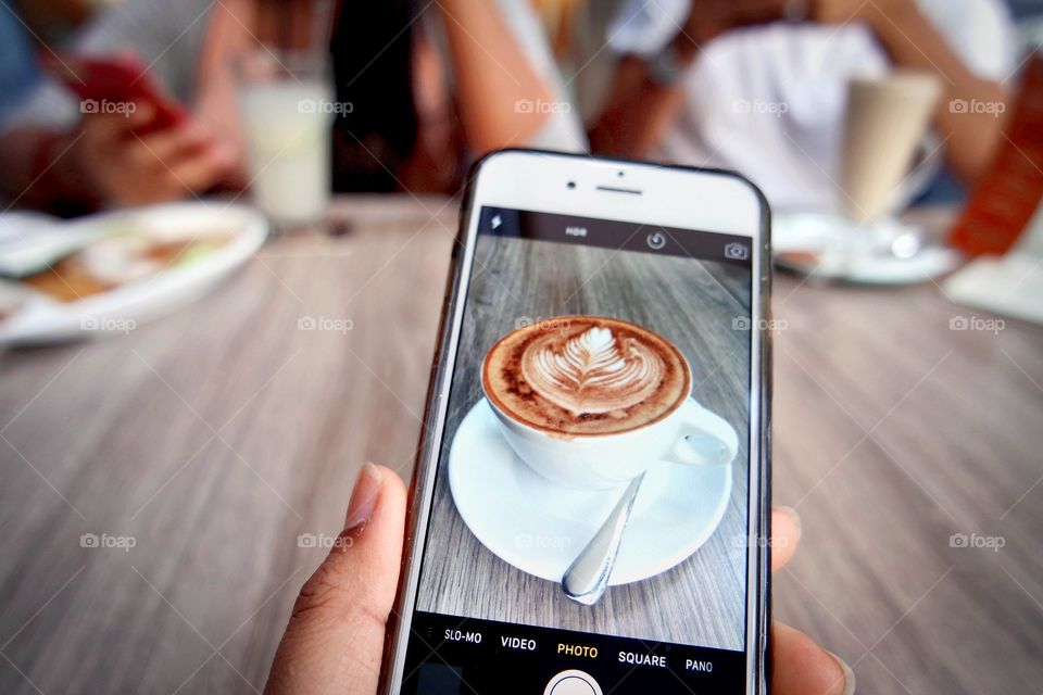 Hand holding smart phone taking a picture of coffee in cafe with friends 