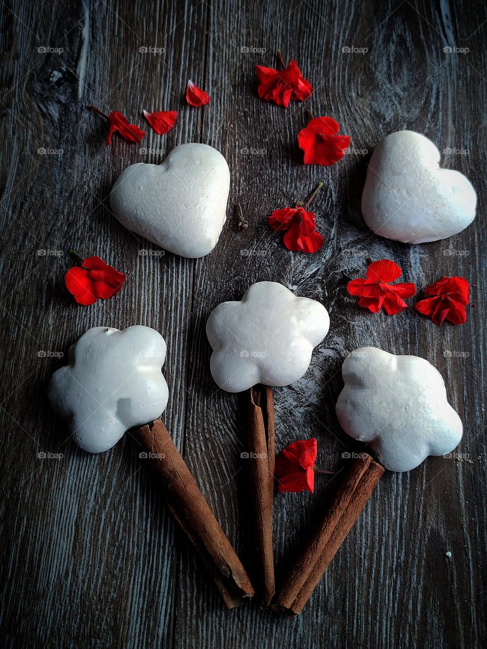 A bouquet of cookies.  On a wooden background are three cinnamon sticks (stems), on top of which are three flower-shaped cookies covered with white icing.  Two heart-shaped cookies covered with white icing lie among red geranium flowers.