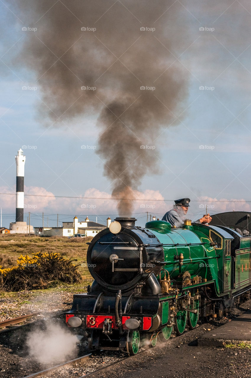 The Romney, Hythe and Dymchurch small railway engine makes a stop near Dungeness lighthouse. There is steam coming from the engine and the driver is onboard