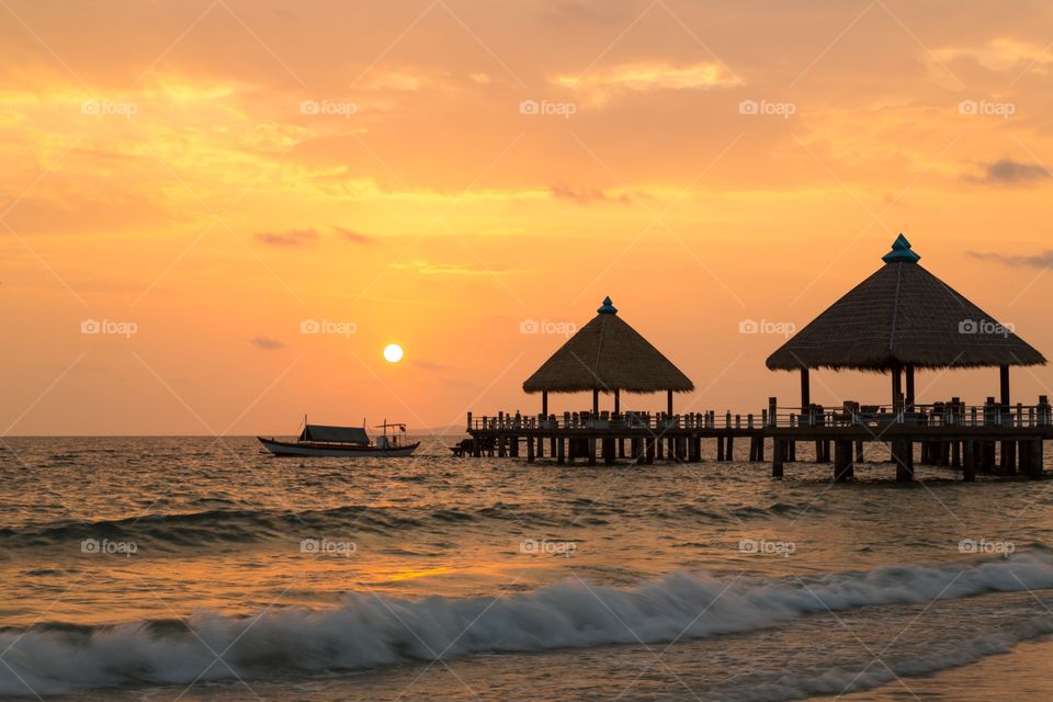 Sunset in Cambodia. Sunset on the beach in Cambodia. Pier in the background with huts and one fishing boat. Orange sky. Waves breaking