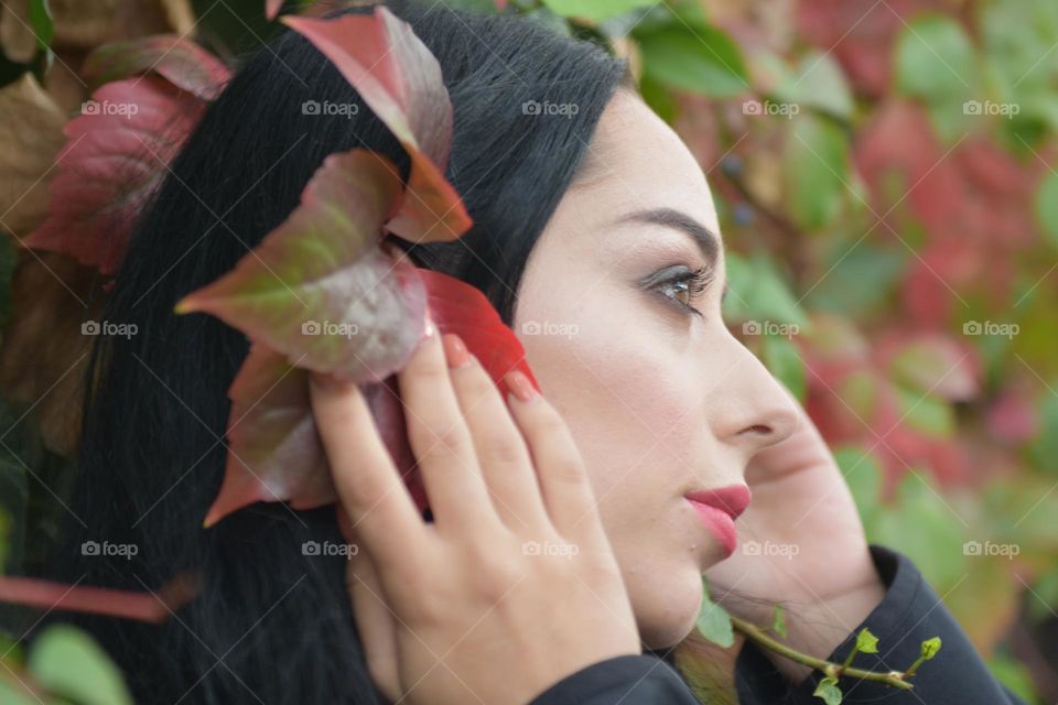 Young fashionable woman in a romantic autumn scenery wearing black coat and fluffy aermuffs with dark green and red leaves in background.Street outdoorfashion. 