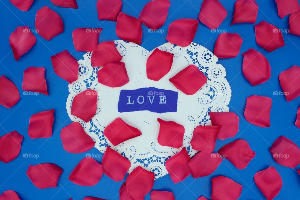 Color Love - The word “LOVE” stamped in white ink on dark blue paper on a heart-shaped white lace doily with scattered bright pink rose petals, all on a bright blue background