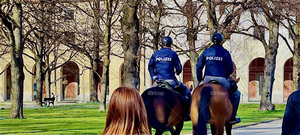 Two horseback policemen are riding through the park adjacent to the “Maximilianeum” in “Munich”, seat of the “Bavarian State Parliament” making their presence known to the people. 2024. Hypnotic Productions