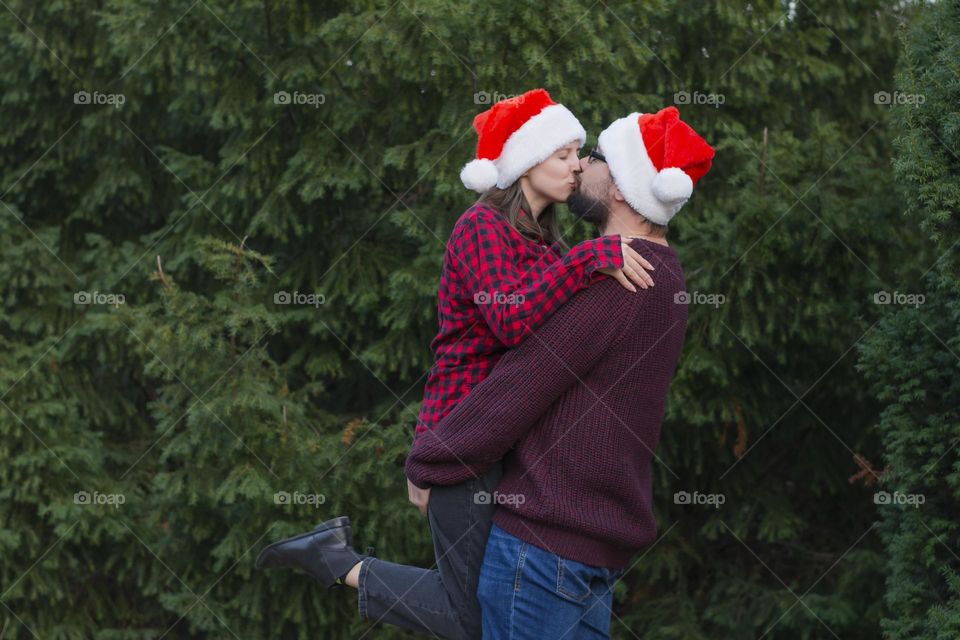 Young beautiful love couple stands on the street near the Christmas trees meets Christmas hugs and kisses in a Santa hat and red sweaters a man raised her in his arms