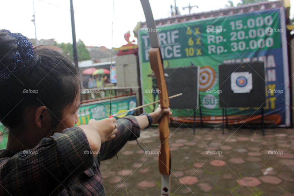 A young Asian woman in plaid shirt holding an archery and aiming at the arrow target in front of her, Semarang, Indonesia.