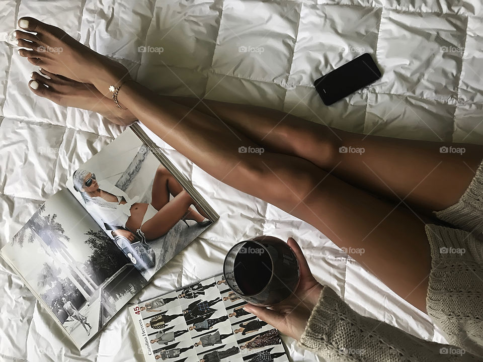 Young woman enjoying wine and reading fashionable magazines in cozy bed 