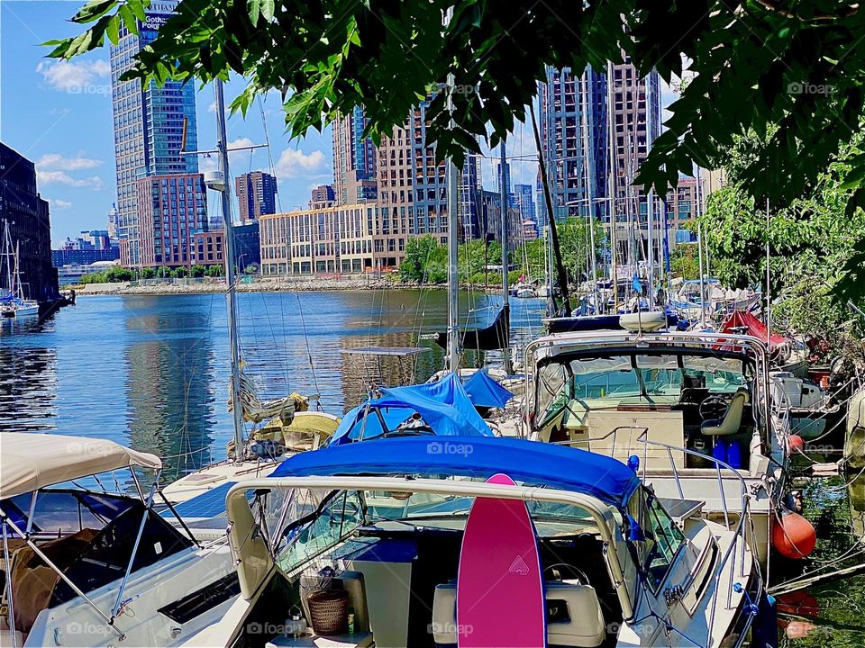 This is a tree lined closeup on the boats at „Newtown Creek“ by the „Pulaski Bridge“ in LIC, Queens. The ocean is featuring silver and gold metallic sparkles and scaffolding encapsulates the construction sites nearby. 2023. Hypnotic Productions