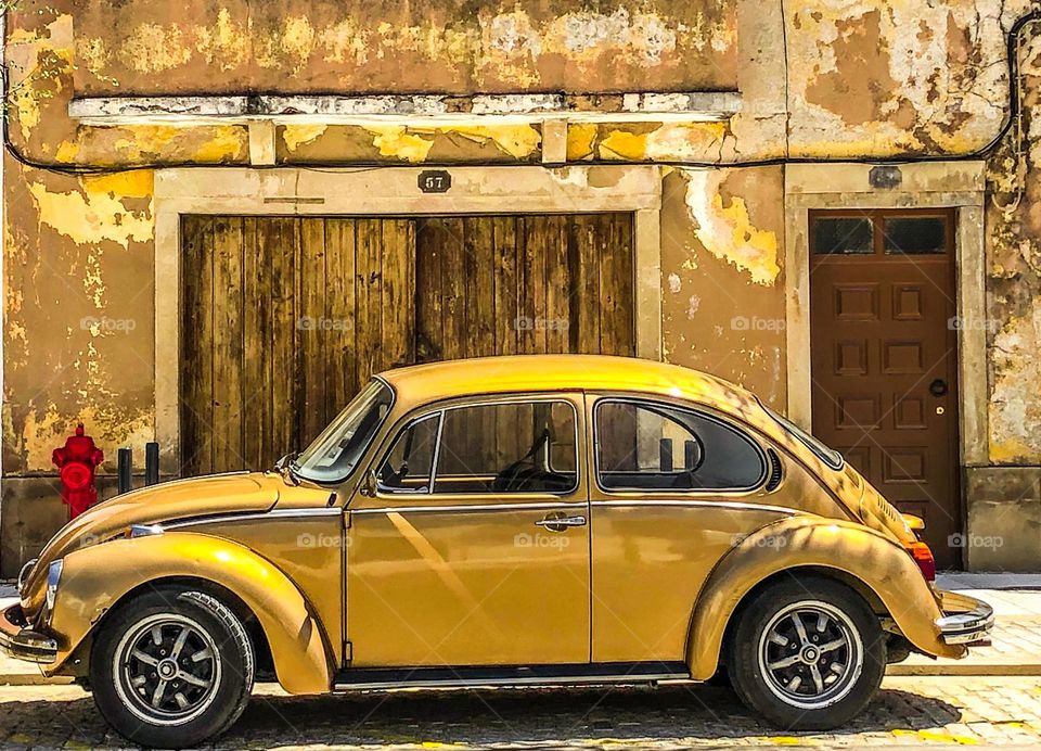 A gold coloured VW Beetle in front of a old building in similar hues, with a red fire hydrant 