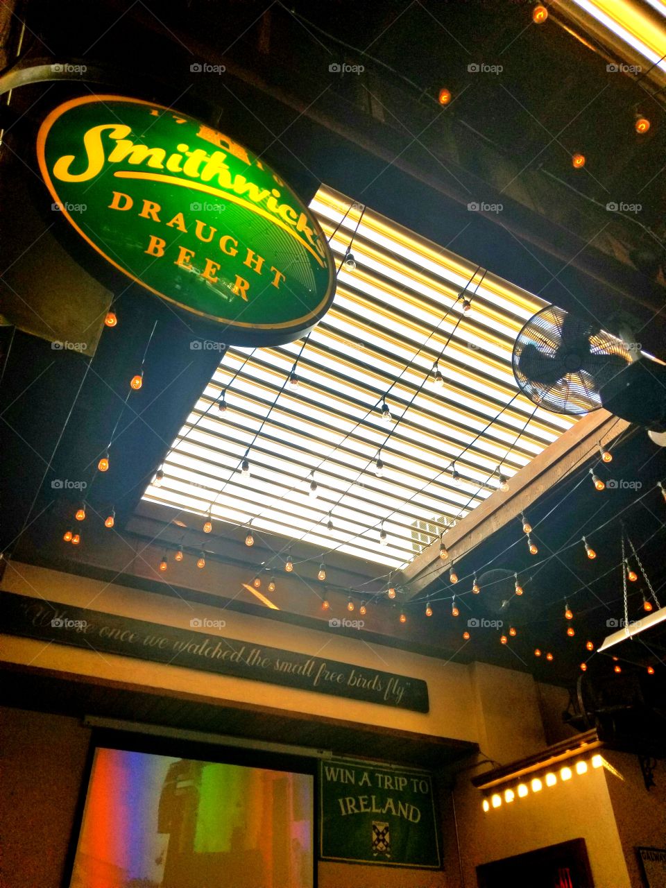 Perfect summer day view at neighborhood pub through open louvered ceiling to blue and white skies decorated with the glow of pub patio bulb string lights and draught beer bar sign