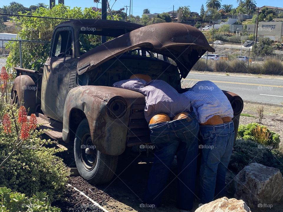 Two men working under the hood of an old rusted out truck as their pumpkin butts show out of the jeans . Very humorous.