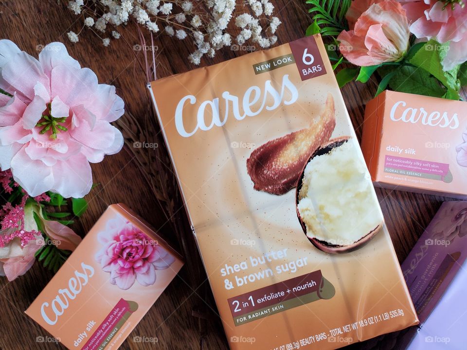The new look Caress bar shea butter and brown sugar six pack centered on a wood surface surrounded by matching florals and two single bars of daily silk on the left and right sides.