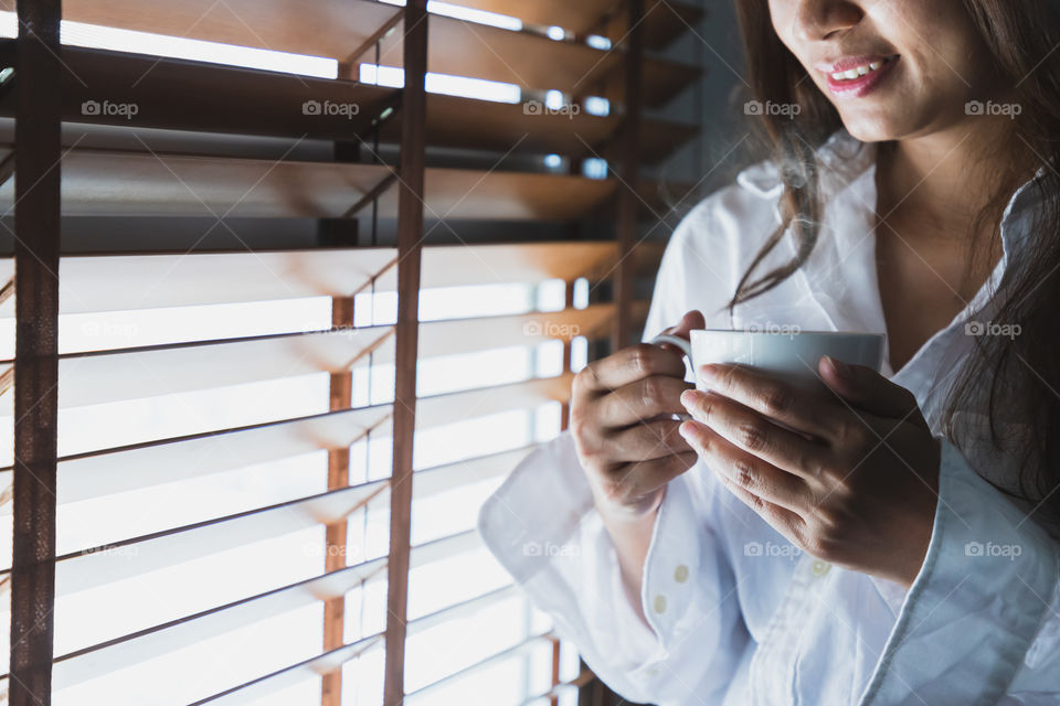 Asian women wearing a white shirt, holding a white coffee mug on the window in the bedroom