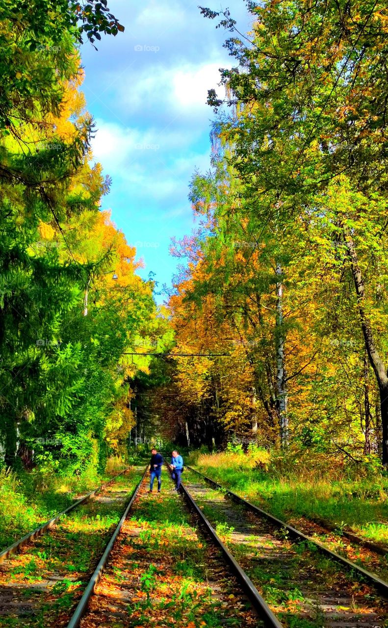 Two men are walking along the tram rails.  Colorful autumn trees along the rails