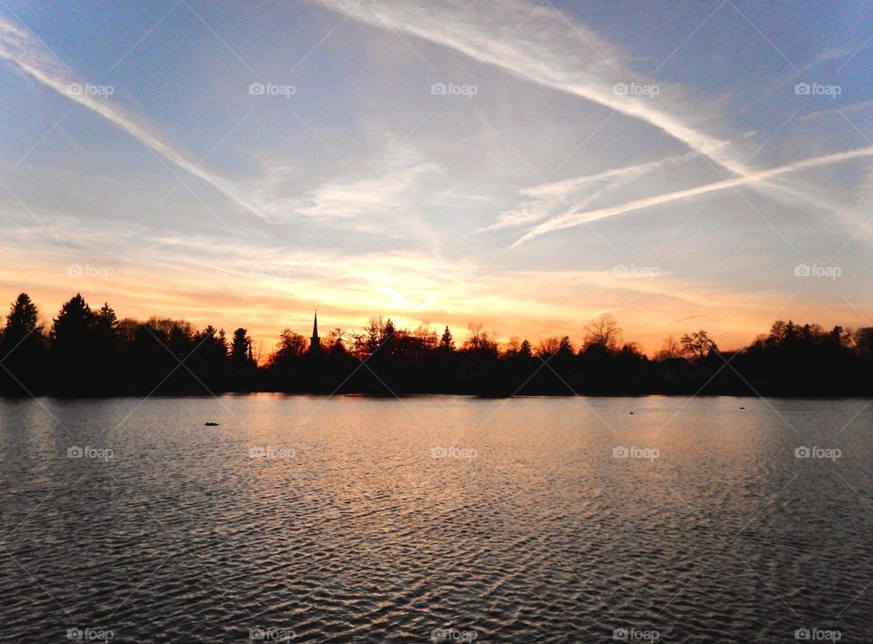 Sunset creates illuminated clouds in orange and white streaks over a midwest Michigan river that reflects the orange colors back to the sky as the trees and church steeple on the riverbank are silhouetted against the sky