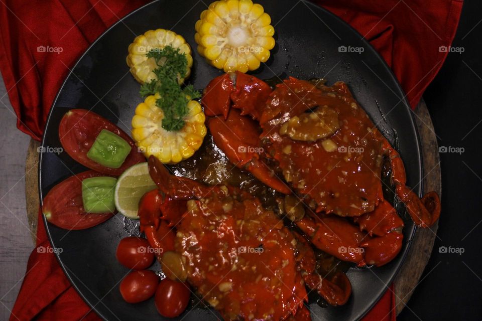 Crab flavored in creamy garlic sauce served with corn, tomatoes, lemon wedges and cherries on a black plate set on a red cloth set in a wooden setting. Asian flavor crab.