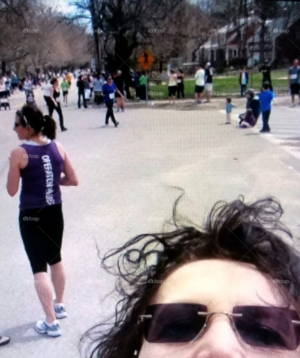 People outside in sunshine, walking or running. Some wearing athletic workout clothes and others in Street clothes. selfie of person taking pic overhead wearing sunglasses 😎 Street treeline & open asphalt area in town square.