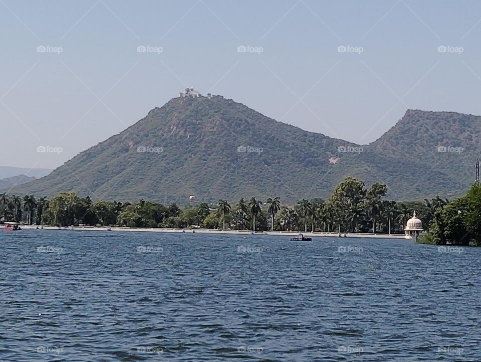 a landscape scene on a river with blue water, mountain, boat, sky, green trees looks very beautiful