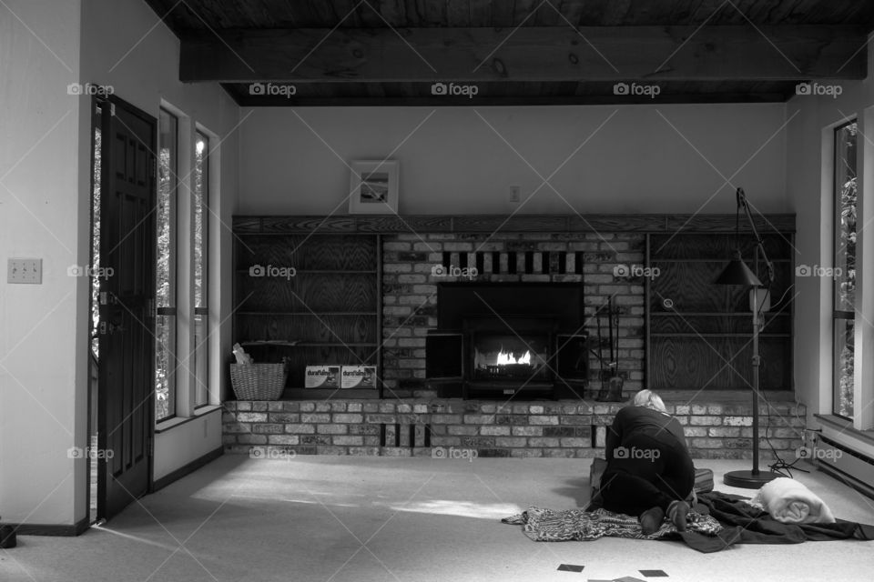 Black and white photo of a fireplace living room, in a cabin in the woods. A woman is laying on the floor facing the fireplace. The room is empty. They are either moving in, moving out, or a squatter squatting in someone else’s empty home.
