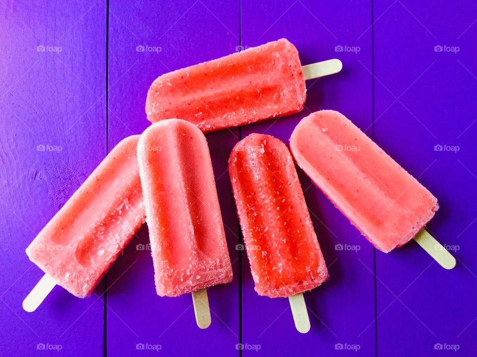 Five delicious, cooling and refreshing, frozen red popsicles with wooden popsicle sticks sitting on a bright purple wooden picnic table.