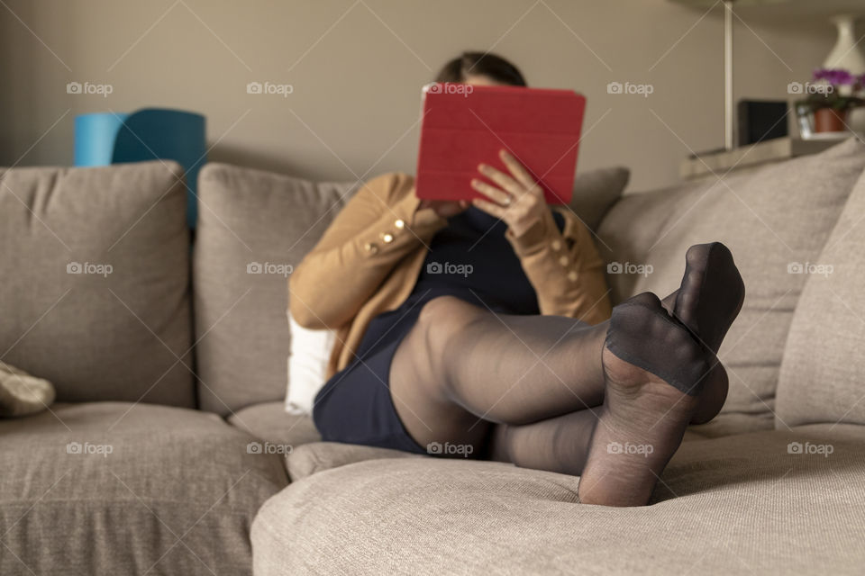 A portrait of a woman sitting in a couch wearing a blue dress and black pantyhose relaxing and looking at her tablet computer.