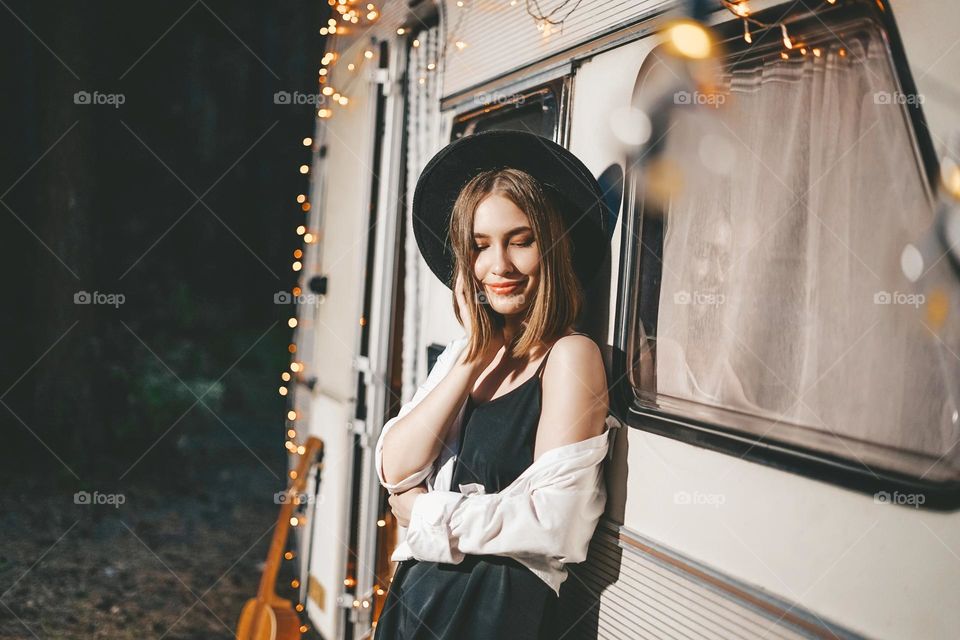 Young beautiful girl in black felt hat and dress in stylish fashionable outfit against background of camper in forest road trip