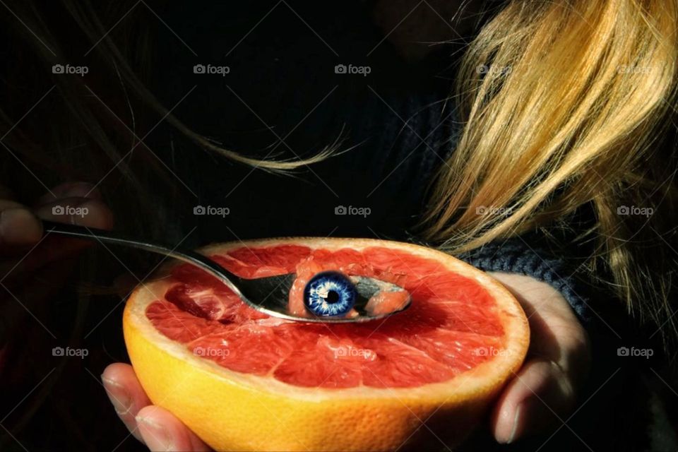 A blue eye lies on a spoon over a pink grapefruit held by a woman with reddish hair