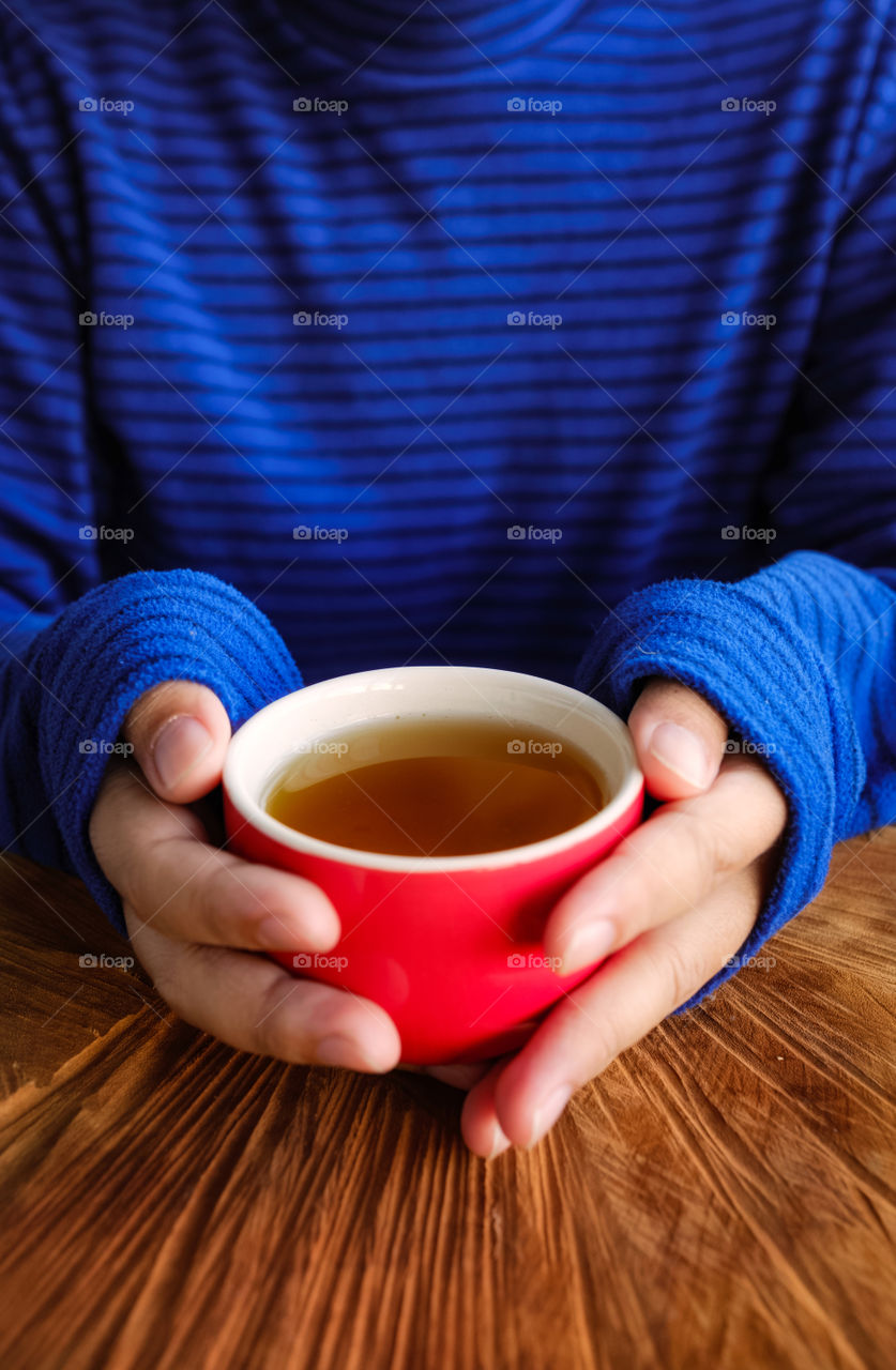 Man hands in a blue sweater holding a red cup of warm tea on the wooden table