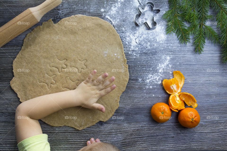 Little child helps cooking christmas ginger cookies on a wooden table with tangerines and green Christmas trees.