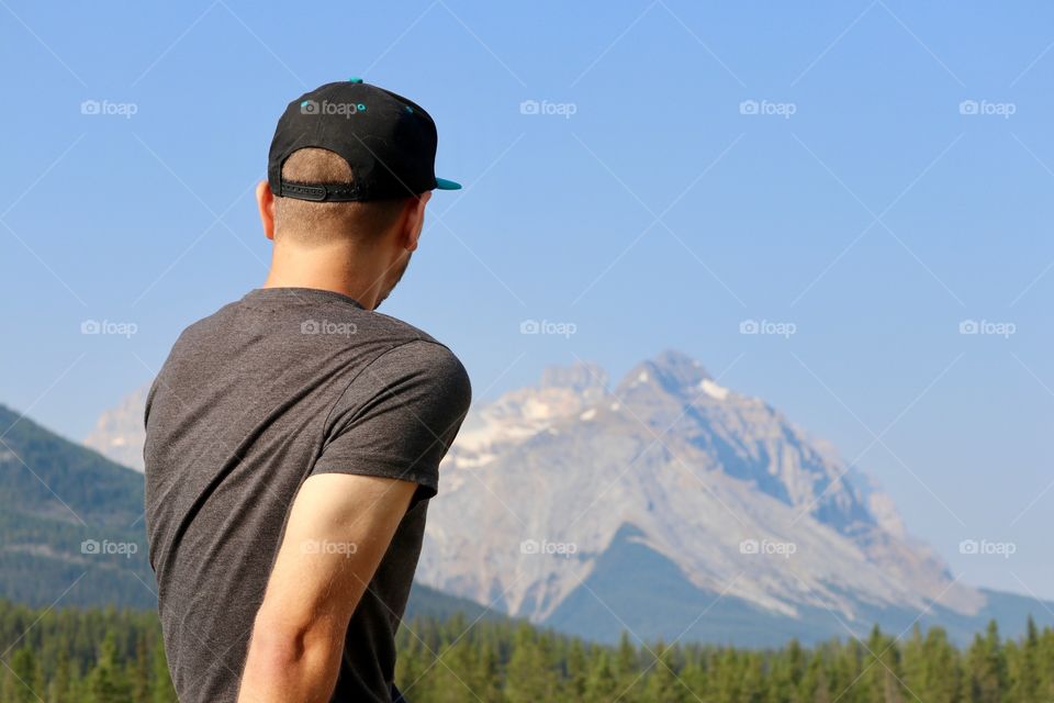 Young man wearing tee shirt and baseball cap viewing Canadian Rocky Mountains from roof of car, space for text 